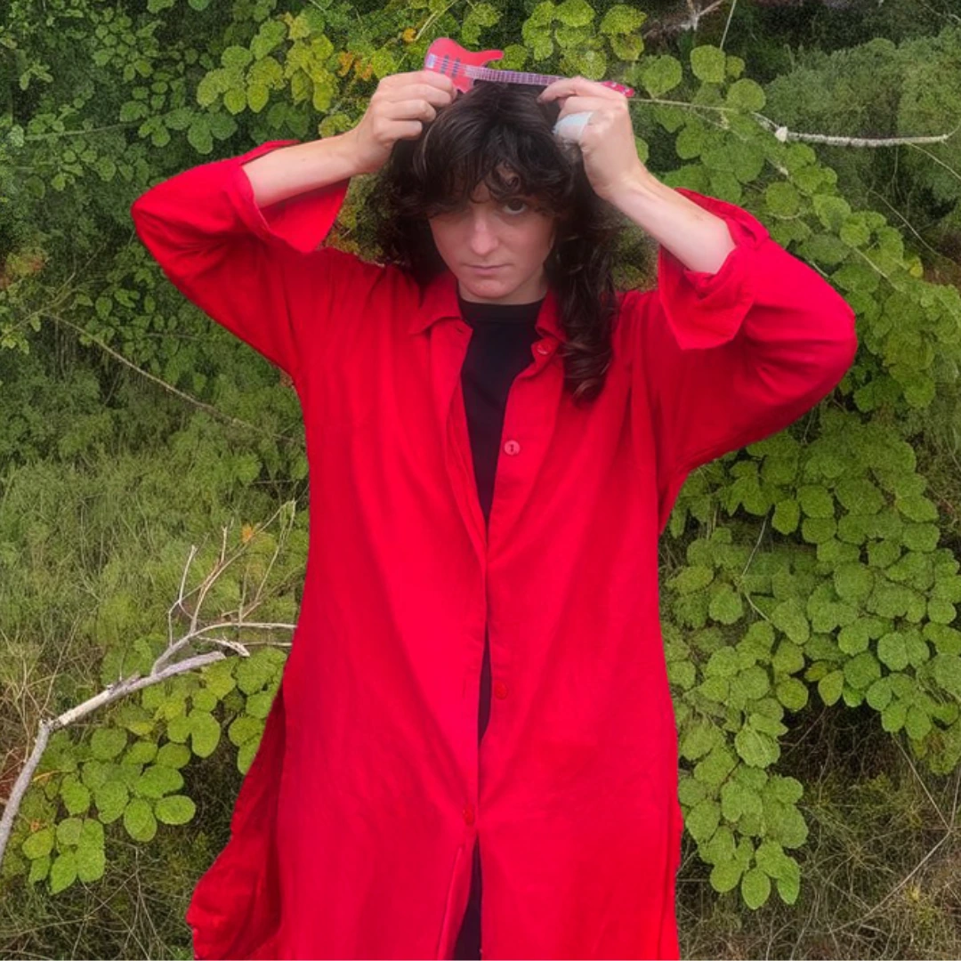 Portrait de Pauline Rouet, membre du duo Blaise & Nico, portant une chemise rouge et tenant une petite guitare au-dessus de sa tête devant un fond de feuillage vert